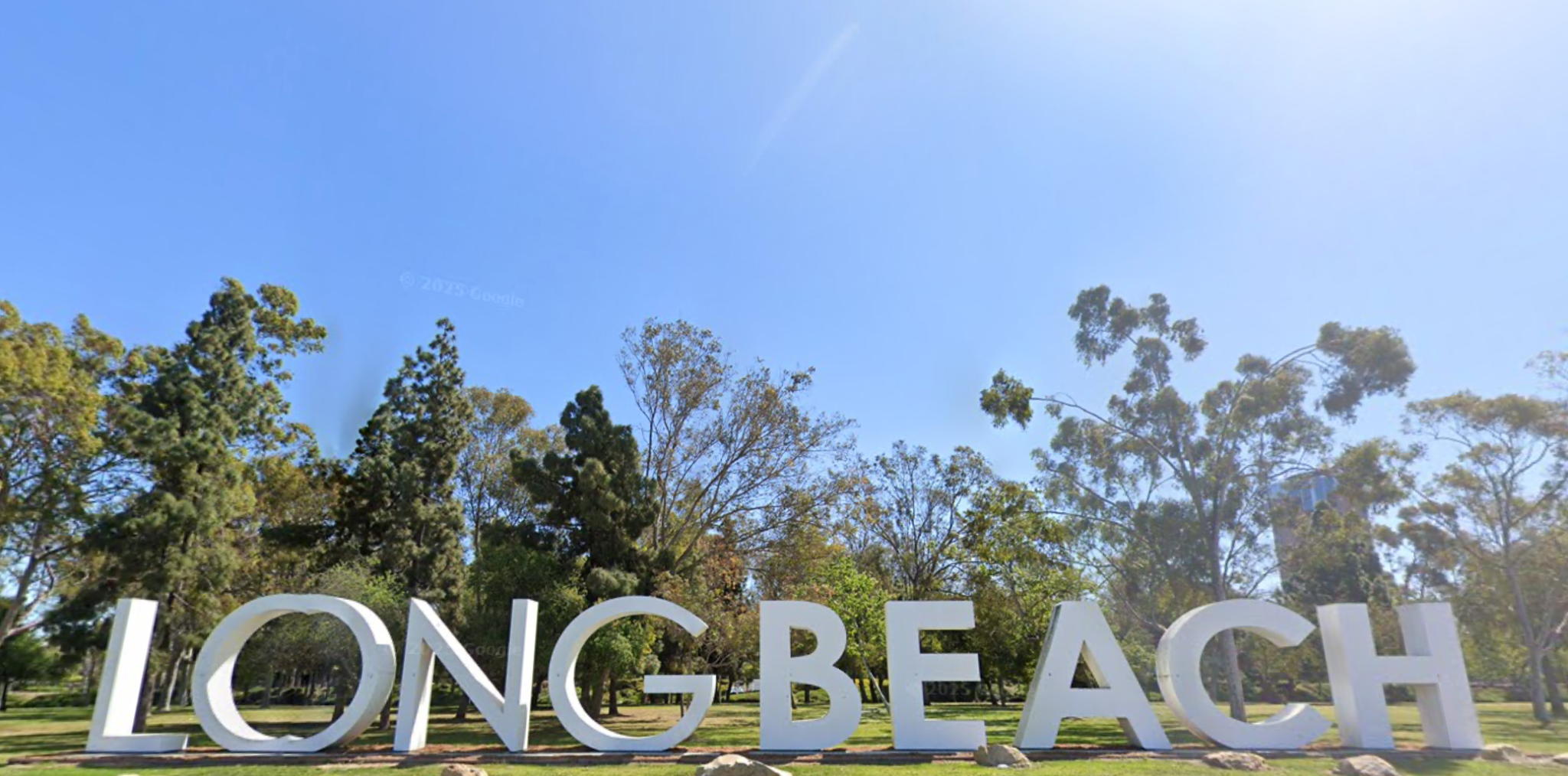 Mature trees behind the large LONG BEACH landmark sculpture, beneath a wide blue sky.